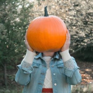 A child holding a Pumpkin by hiding her face with it.